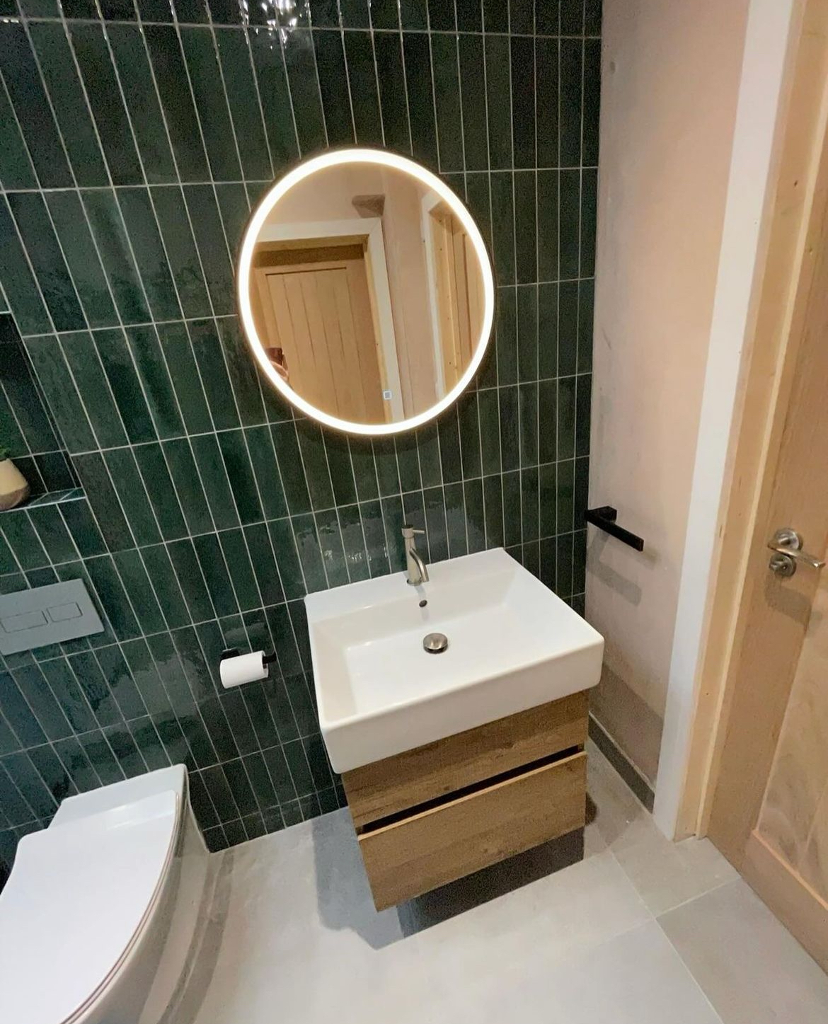 Green tiled cloakroom with oak vanity unit, basin and illuminated round mirror
